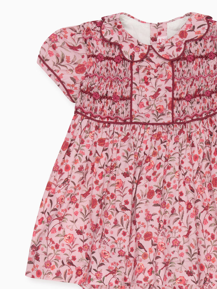 A pink short-sleeve baby dress with a red and green floral print, featuring smocked bodice details and a ruffled Peter Pan collar.