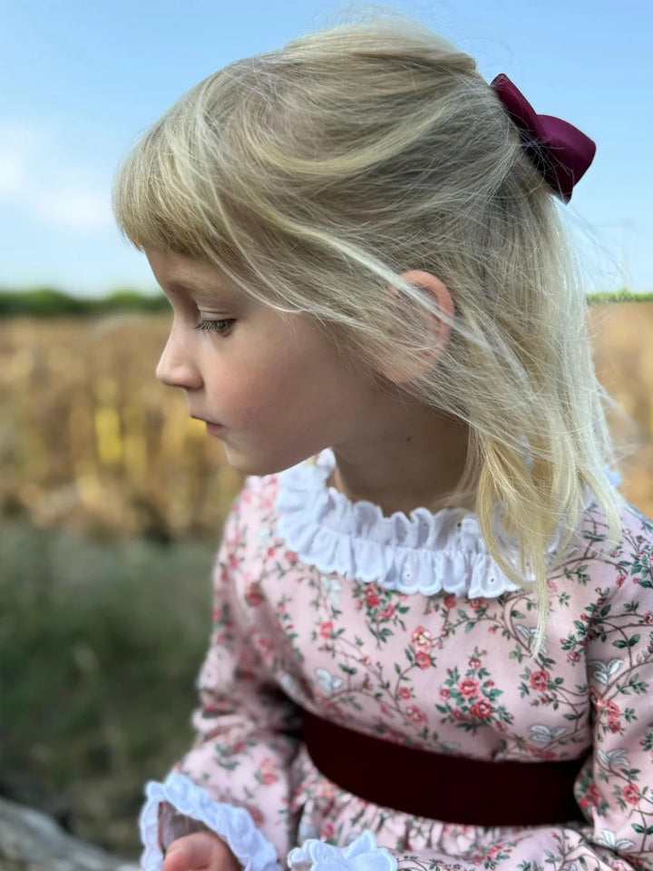A young girl with blonde hair tied back with a dark red bow, wearing a pink floral dress with a white ruffled collar and a dark red sash, photographed outdoors against a blurred natural background.