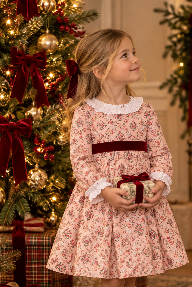 A young girl wearing a pink floral long-sleeve dress with a dark red waist sash and white ruffled collar, holding a wrapped gift box in front of a decorated Christmas tree.