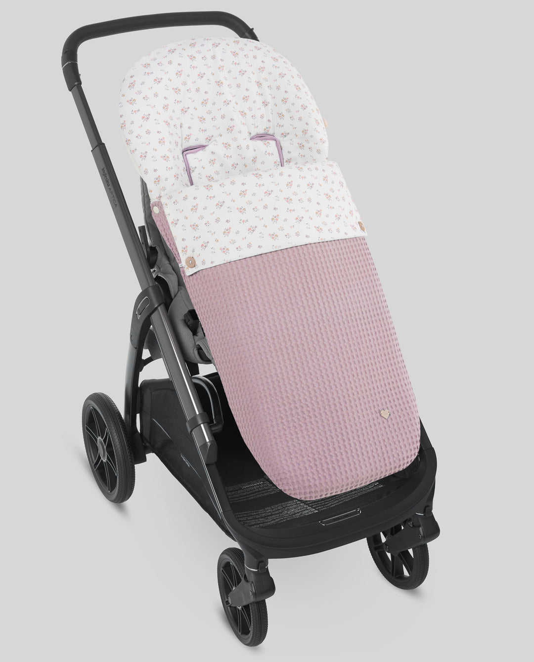 A stroller footmuff and seat cover set in a black stroller, featuring a white top section with a tiny pink floral pattern and a dusty pink waffle-textured lower section.
