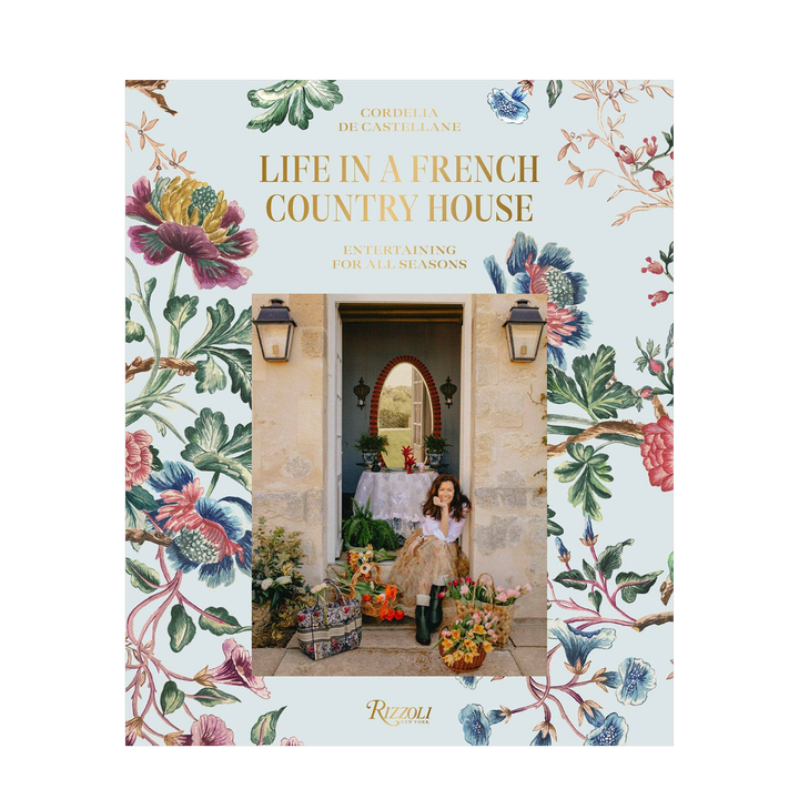The front cover of the book 'Life in a French Country House' by Cordelia de Castellane, published by Rizzoli. The cover features a light blue background decorated with vintage floral illustrations on the borders, framing a central photograph of a young girl in a vintage dress standing in a stone courtyard doorway.