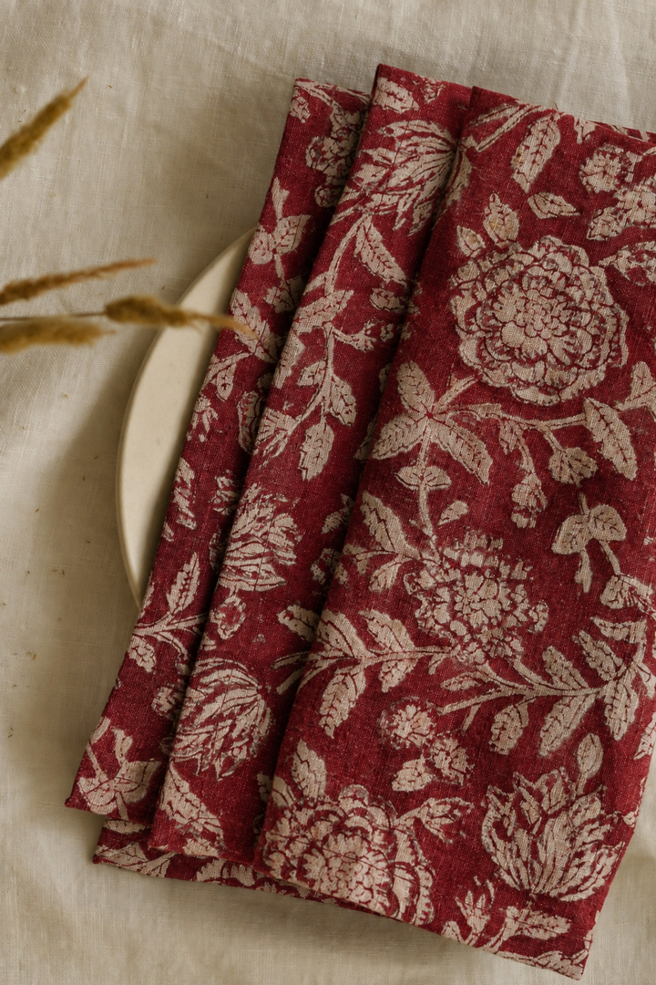 A folded red fabric napkin with a beige floral block-print pattern, styled on a textured beige linen surface next to a plate and dried grass.