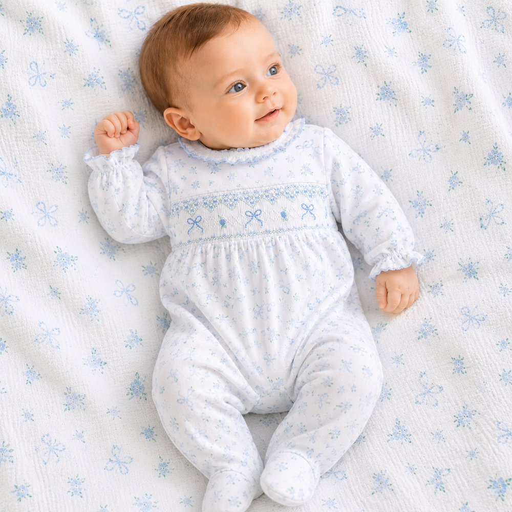 Baby lying on a white blanket with blue snowflake patterns, wearing a white outfit with blue floral embroidery.