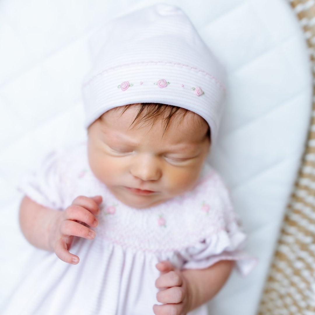 Newborn baby wearing a white hat and outfit with pink details, sleeping peacefully.