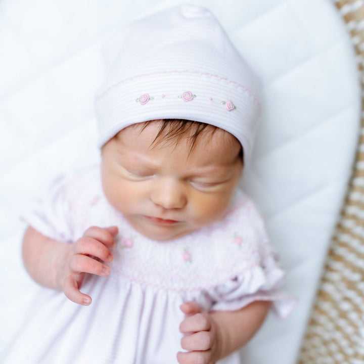 Newborn baby wearing a white hat and outfit with pink details, sleeping peacefully.