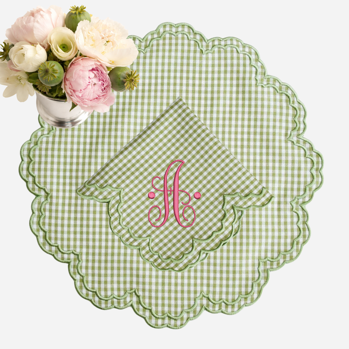 Green gingham placemat with scalloped edge and pink monogram, accompanied by a floral arrangement.