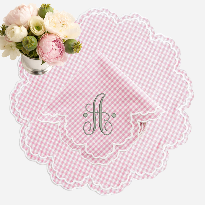 Pink gingham placemat with scalloped edge and monogram 'A' a pink gingham napkin on a white background next to a floral arrangement
