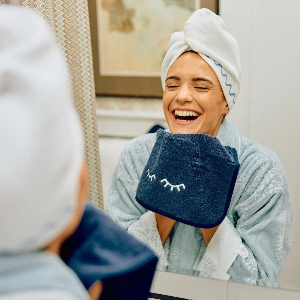 Woman in a robe with a towel on her head, holding a blue face mask with a smiley face design.