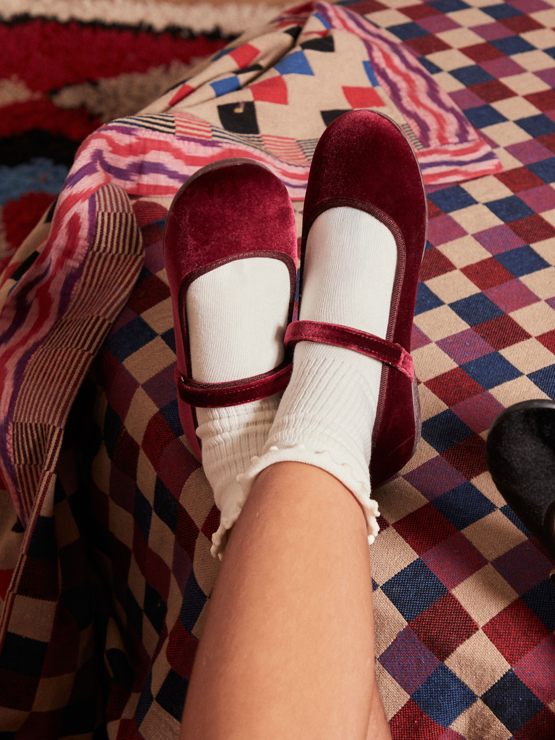 A person's feet wearing burgundy velvet Mary Jane shoes with white ribbed socks, resting on a patterned red, blue, and beige plaid blanket.