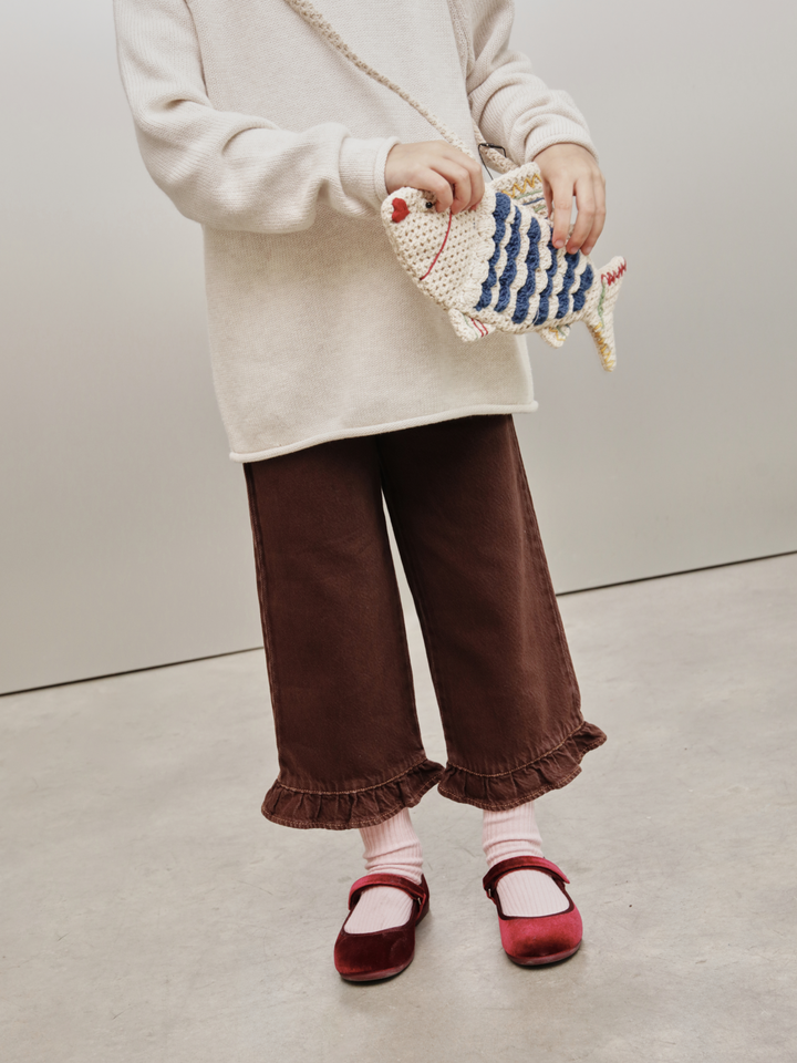 A child wearing a cream knit sweater, brown wide-leg corduroy pants with ruffled hems, pink socks, and burgundy Mary Jane shoes, holding a small crocheted fish-shaped clutch bag.