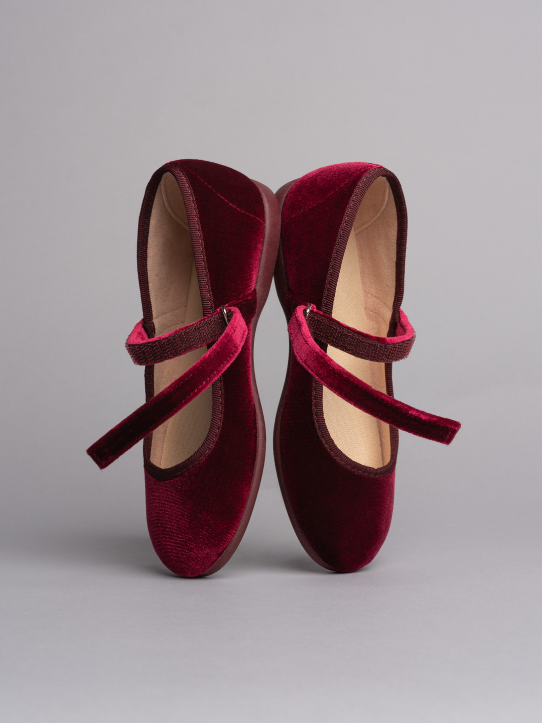 A pair of deep burgundy velvet ballet flats with matching velvet straps, displayed against a neutral gray background.