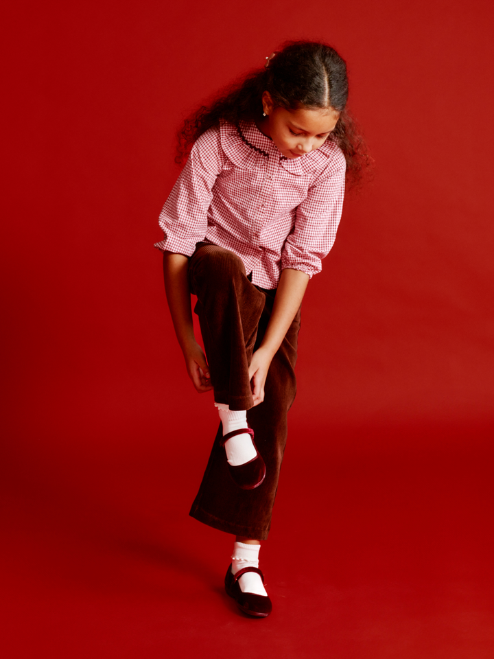 A young girl with curly hair is shown from the waist down, looking down as she adjusts her brown pants. She is wearing a pink and white gingham button-down shirt, brown corduroy pants, white socks, and dark brown Mary Jane shoes, set against a solid red studio background.