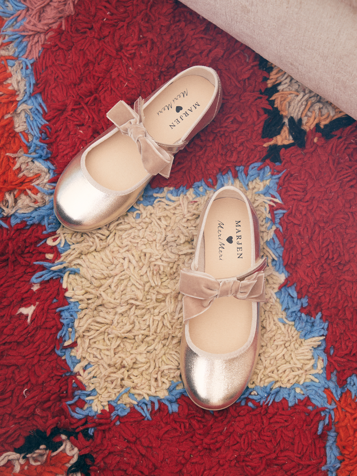 A pair of metallic gold Mary Jane shoes with beige satin bows, displayed on a colorful shag rug. The brand name 'MARGIE' is visible on the insoles.