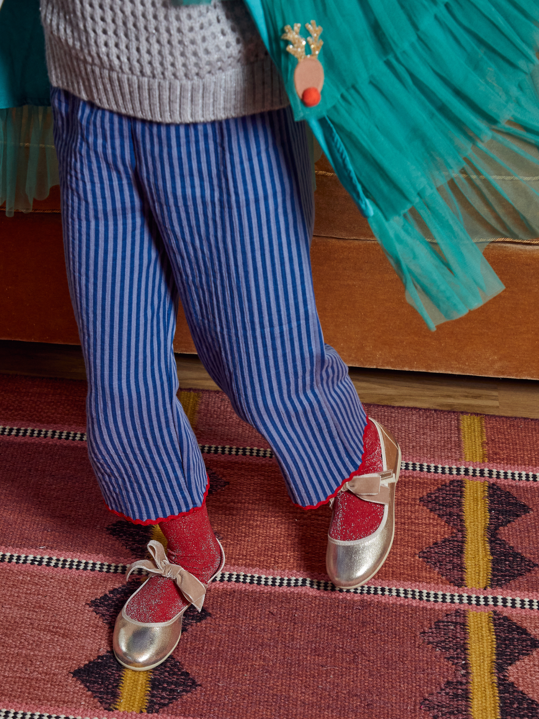 A child's lower body wearing blue and white vertically striped pants, red knit socks, shiny silver ballet flats with bow details, and a gray knit sweater. A turquoise tulle skirt or cape is draped over the shoulder, and the child stands on a patterned rug.