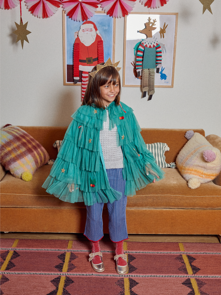 A young girl stands in a decorated living room wearing a bright teal tulle cape with ruffled layers and small red berry details, paired with blue-and-white striped pants, red socks, and red-and-gold shoes. A brown sofa, patterned pillows, framed wall art, and hanging paper decorations are visible in the background.