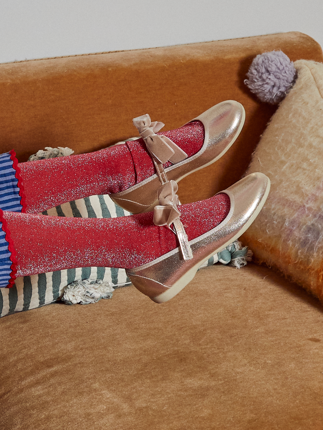A pair of metallic rose gold Mary Jane shoes with bow straps, worn with red glittery socks, resting on a brown velvet couch with striped pillows.