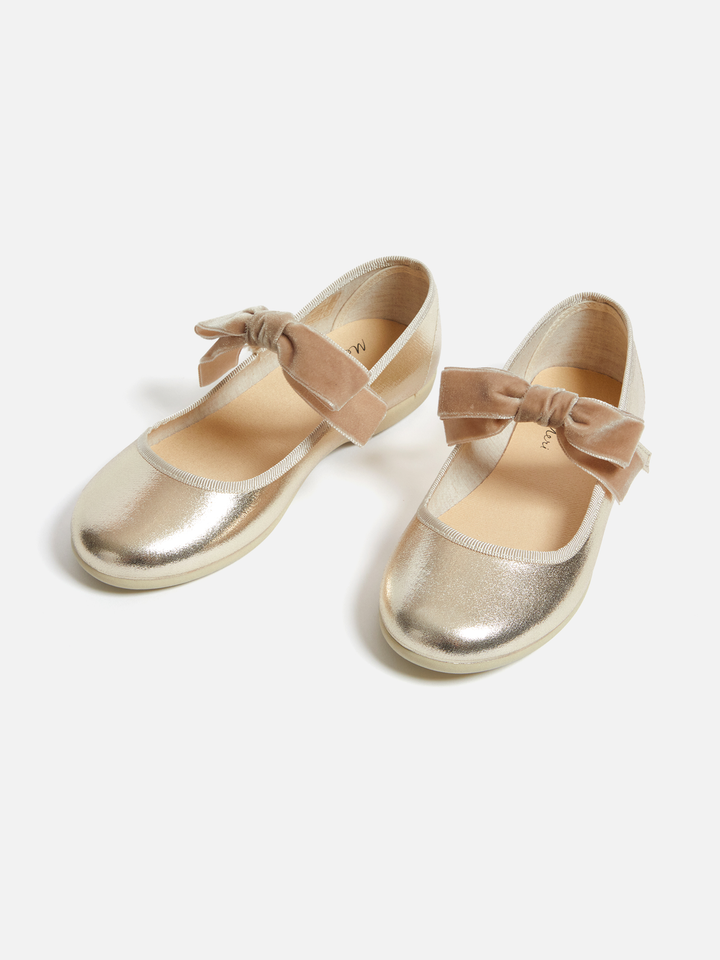A pair of metallic gold ballet flats with a beige velvet bow on the toe strap, shown on a light gray background.