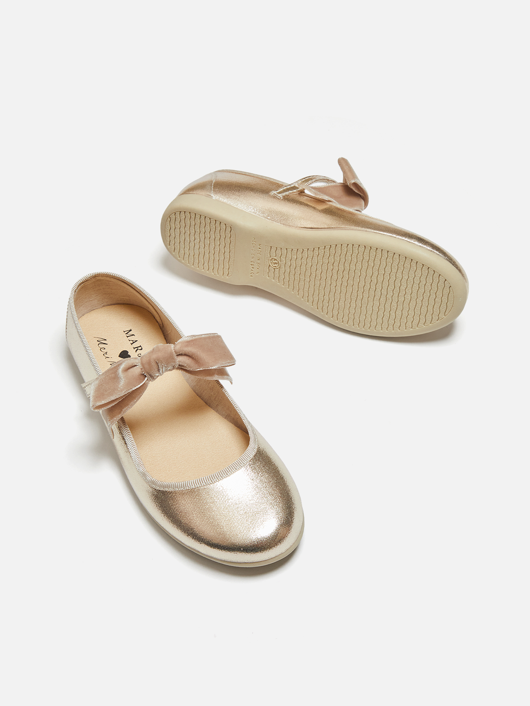 A pair of metallic gold Mary Jane flats with a dusty pink velvet bow, shown on a clean white background. One shoe is oriented to show the side profile with the bow, and the other is turned to show the sole.