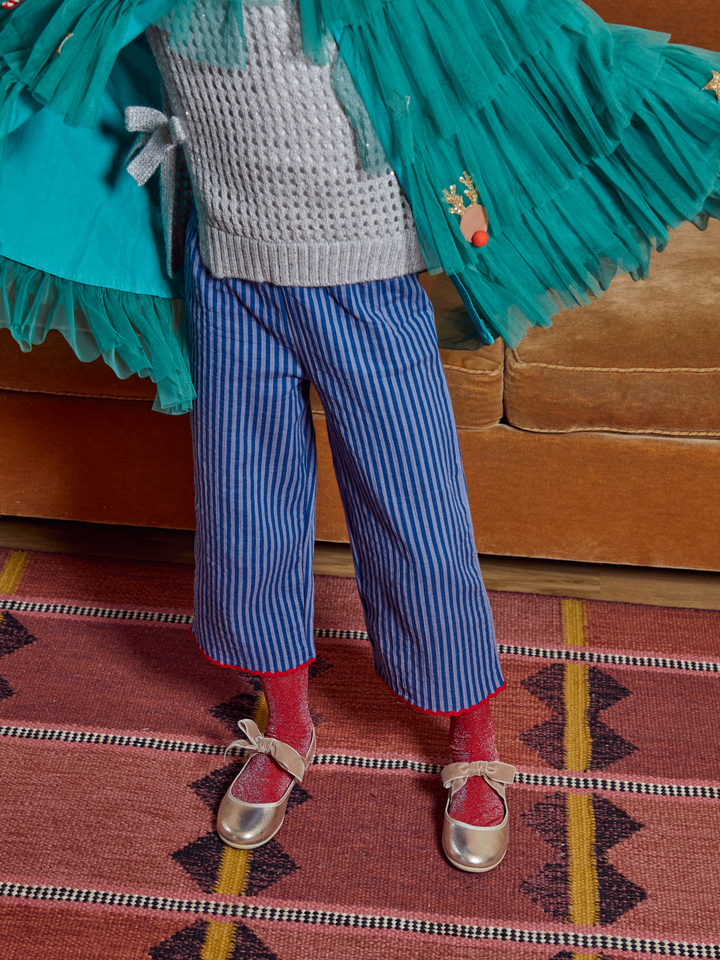 A child wearing blue and white vertically striped pants, red socks, and metallic gold shoes, styled with a gray knit sweater and a large teal tulle tutu overlay, standing on a patterned rug in front of a brown couch.