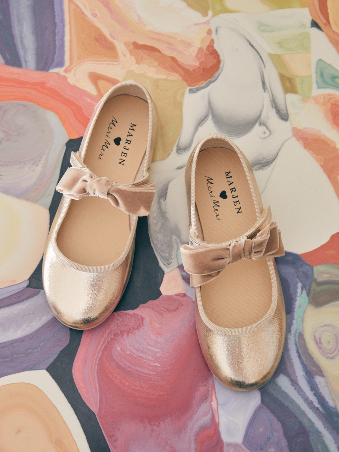 A pair of metallic gold Mary Jane shoes with dusty pink velvet bows, displayed on a colorful abstract painted background. The brand name 'MARJEN' is visible on the insoles.