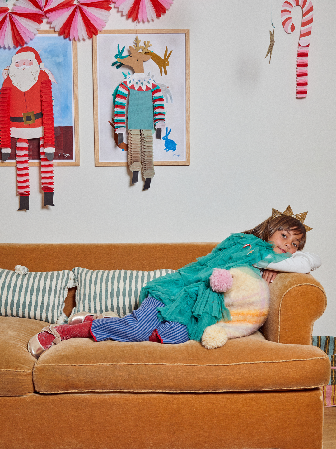 A child lies on an orange-brown velvet sofa in a decorated living room, wrapped in a teal knit blanket and wearing blue-and-red striped socks and tights. Framed handmade holiday artwork hangs on the wall above the couch.