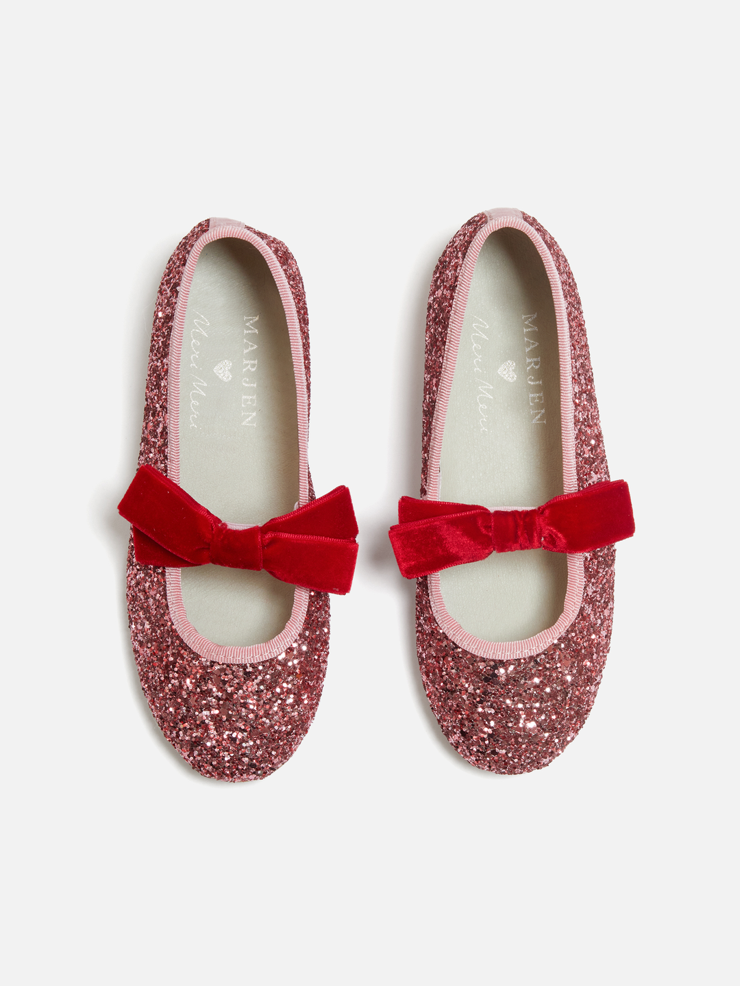 A pair of children's Mary Jane flats by Marjolin, covered in sparkling dusty rose glitter with a vibrant red velvet bow on the toe, shown from a top-down view on a light gray background.