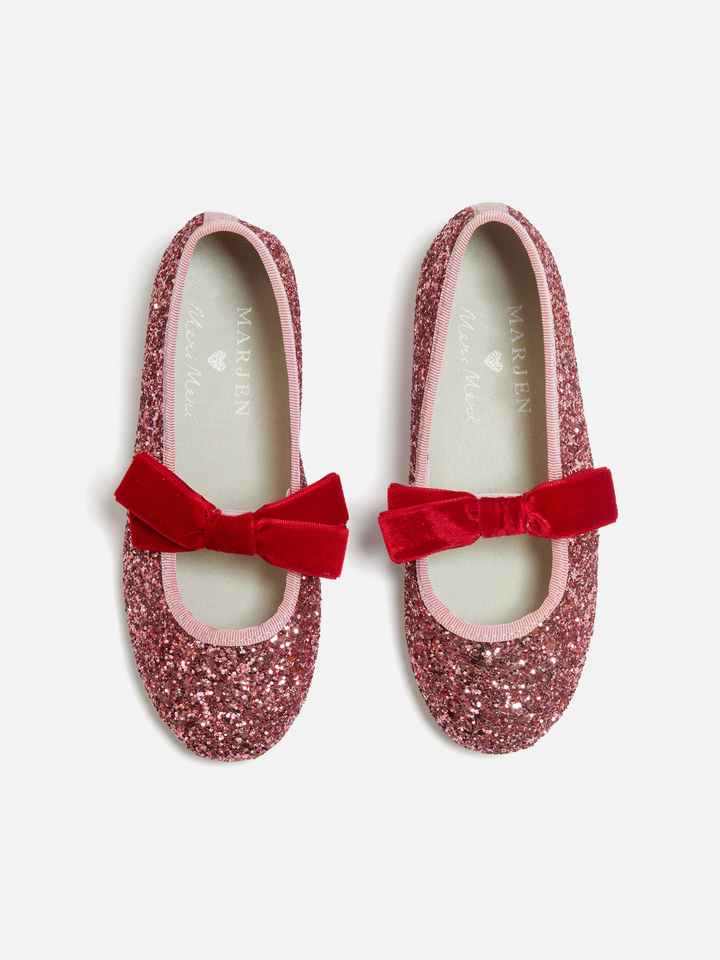 A pair of children's Mary Jane flats by Marjolin, covered in sparkling dusty rose glitter with a vibrant red velvet bow on the toe, shown from a top-down view on a light gray background.