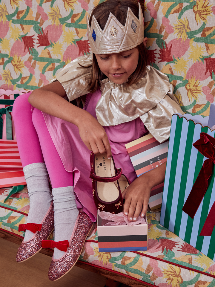 A young girl dressed as a princess sits on a floral bench surrounded by gift bags, wearing a shiny gold tiara and a pink dress while trying on a pair of burgundy Mary Jane shoes in an open box.