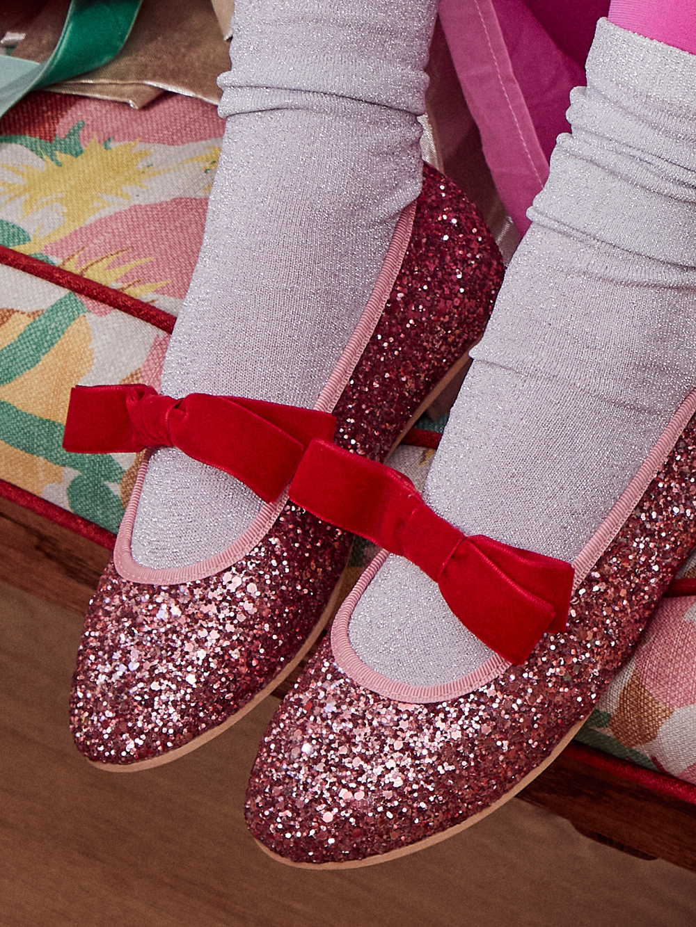 A close-up of a person's feet wearing sparkly pink Mary Jane shoes with red velvet bows and light gray ribbed socks, sitting on a patterned bench.