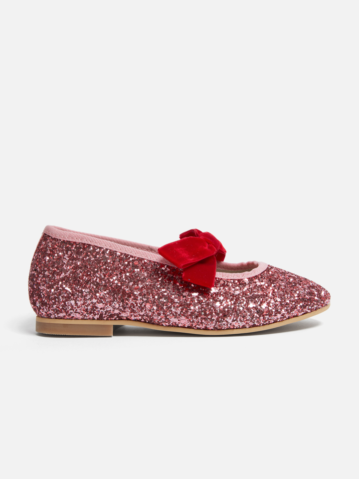 A single pink glitter flat shoe with a red velvet bow on the toe, shown in profile against a light gray background.