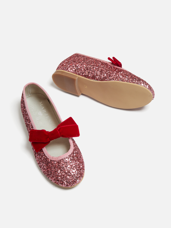 A pair of girls' glitter flats in pink with a red velvet bow on the front and a small red tassel detail on the side, shown on a light gray background.