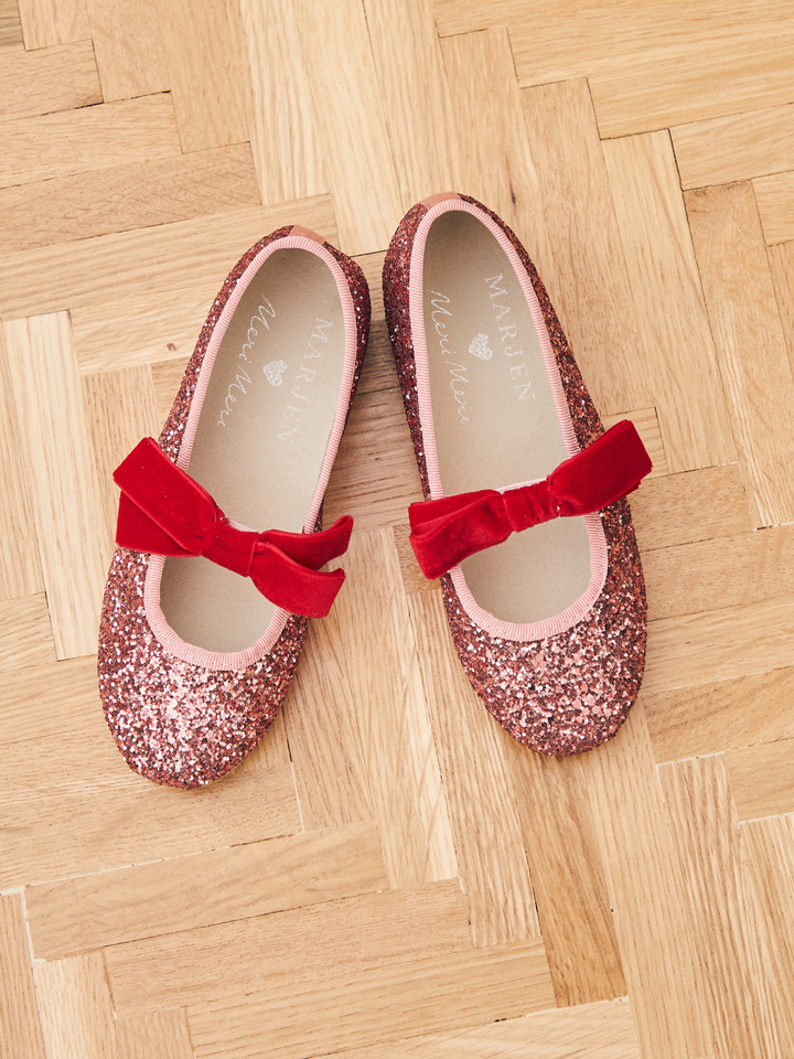 A pair of children's Mary Jane flats with pink glitter uppers and red velvet bows, displayed on a light wood parquet floor. The brand name 'Alice May' is printed on the insoles.