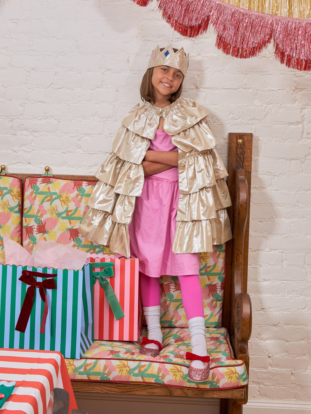 A young girl wearing a pink princess dress, shiny gold cape, and matching gold crown sits on a floral bench with gift bags and a festive red fringe banner in the background.