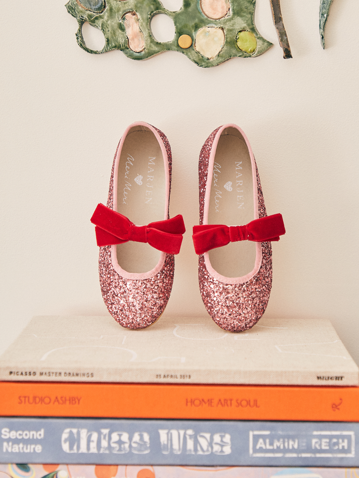 A pair of pink glitter Mary Jane shoes with red velvet bow accents, displayed on a stack of books against a cream wall.