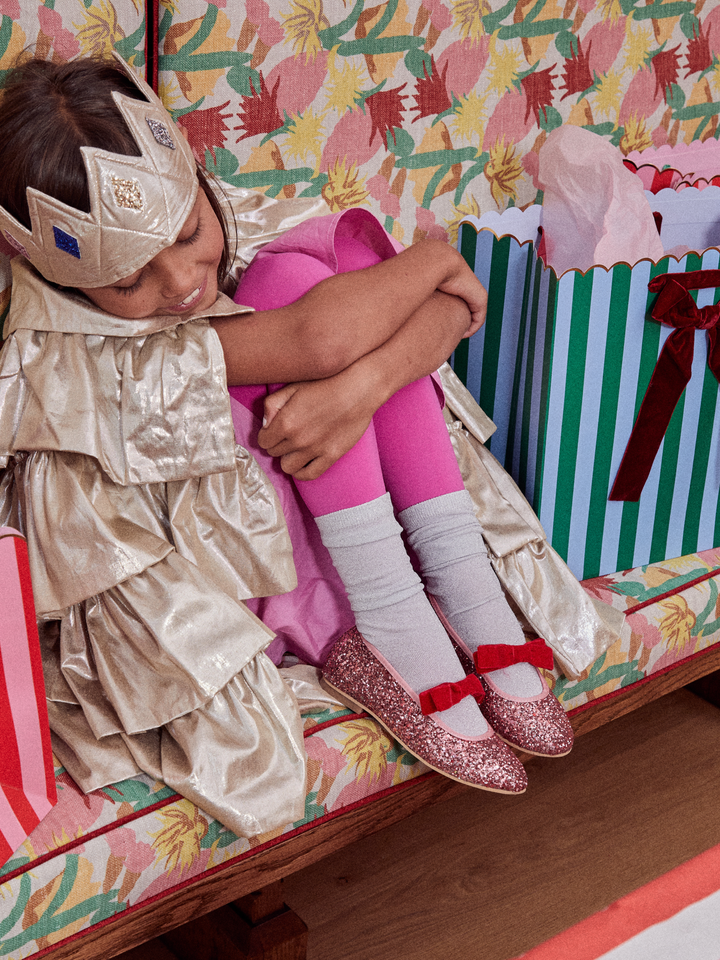 A young girl wearing a shiny gold princess costume, pink tights, and sparkly pink shoes sits hugging her knees on a patterned bench with colorful gift bags in the background.
