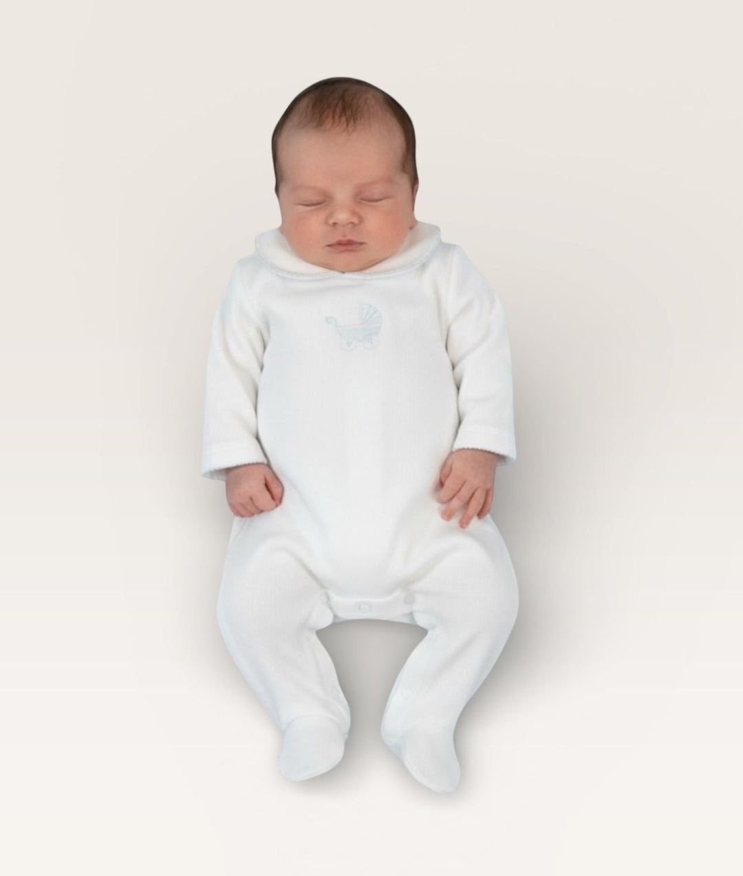 A sleeping newborn baby wearing a white long-sleeve footed sleeper, photographed from a top-down view against a solid off-white background.