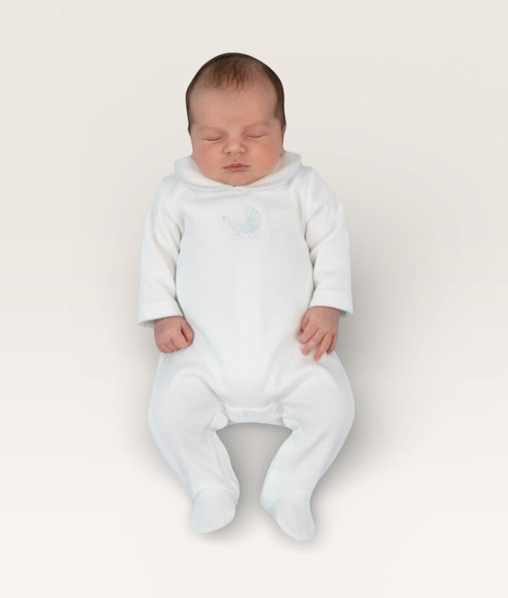 A sleeping newborn baby wearing a white long-sleeve footed sleeper, photographed from a top-down view against a solid off-white background.