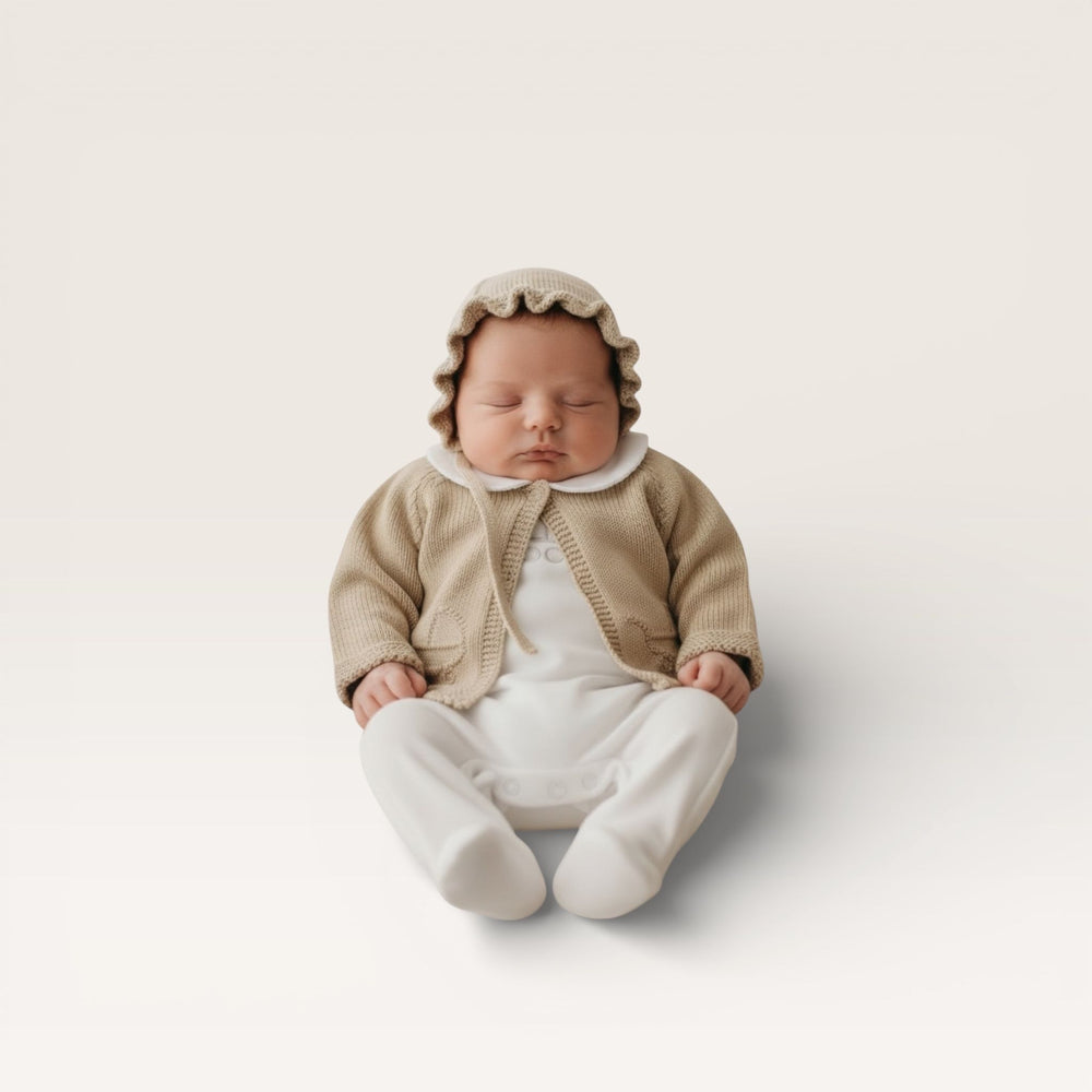 A sleeping newborn baby wearing a beige knit bonnet, cardigan, and white footed romper, sitting against a clean, light beige studio background.