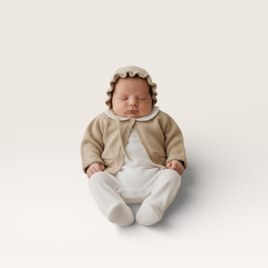 A sleeping newborn baby wearing a beige knit bonnet, cardigan, and white footed romper, sitting against a clean, light beige studio background.