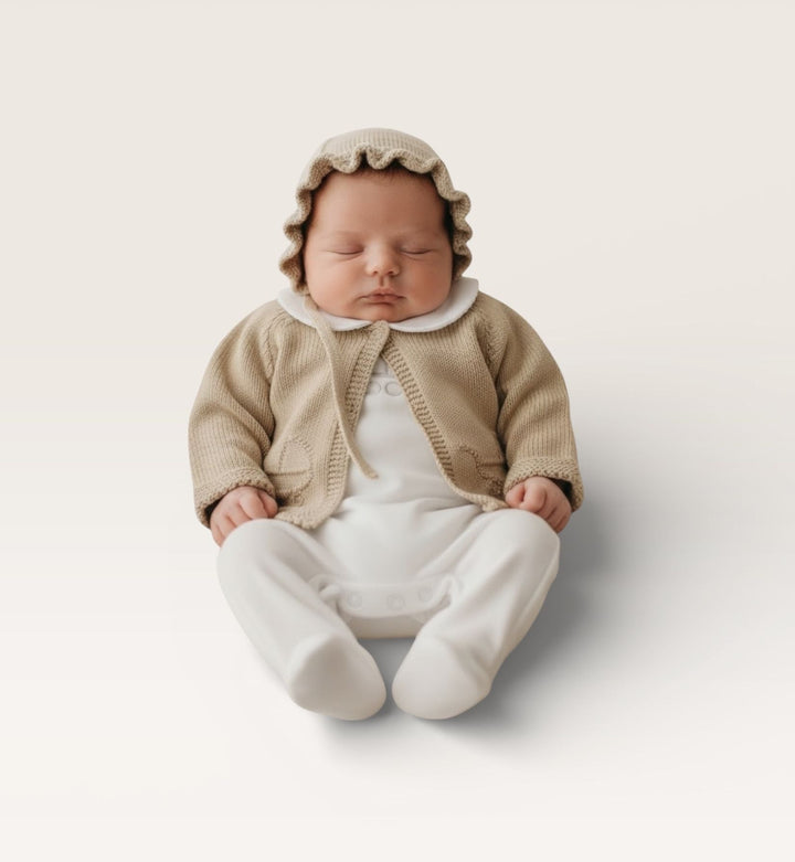 A sleeping newborn baby wearing a beige knit bonnet, cardigan, and white footed romper, sitting against a clean, light beige studio background.
