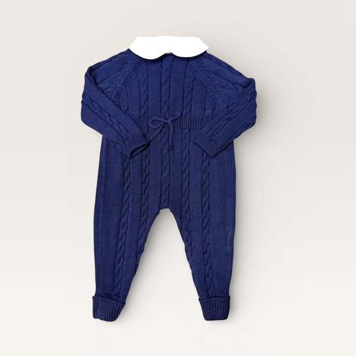 A baby cable-knit jumpsuit in deep navy blue, featuring a contrasting white Peter Pan collar and a small drawstring detail at the chest, laid flat against a light beige background.