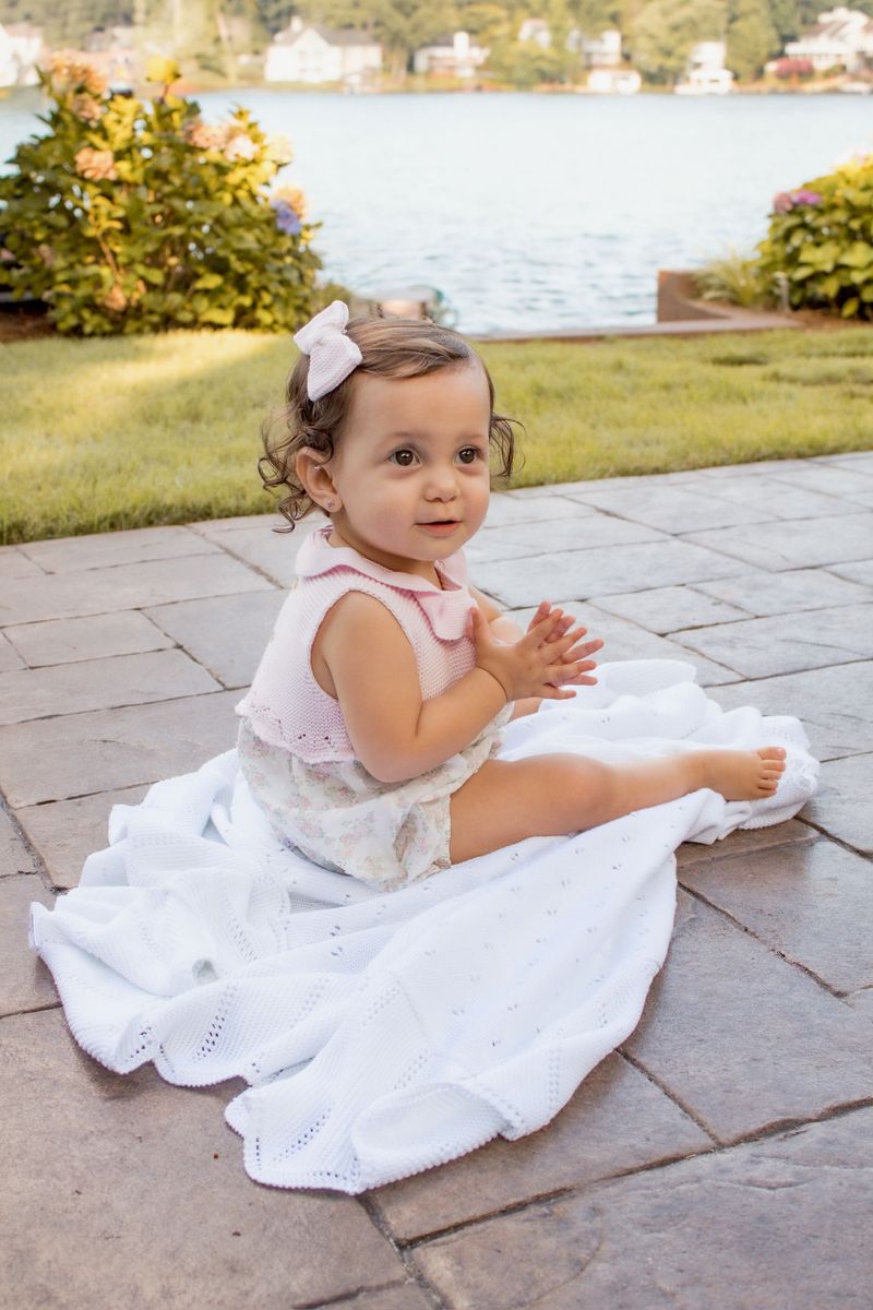 Child in a white outfit with a pink bow sitting on a stone patio by a lake.