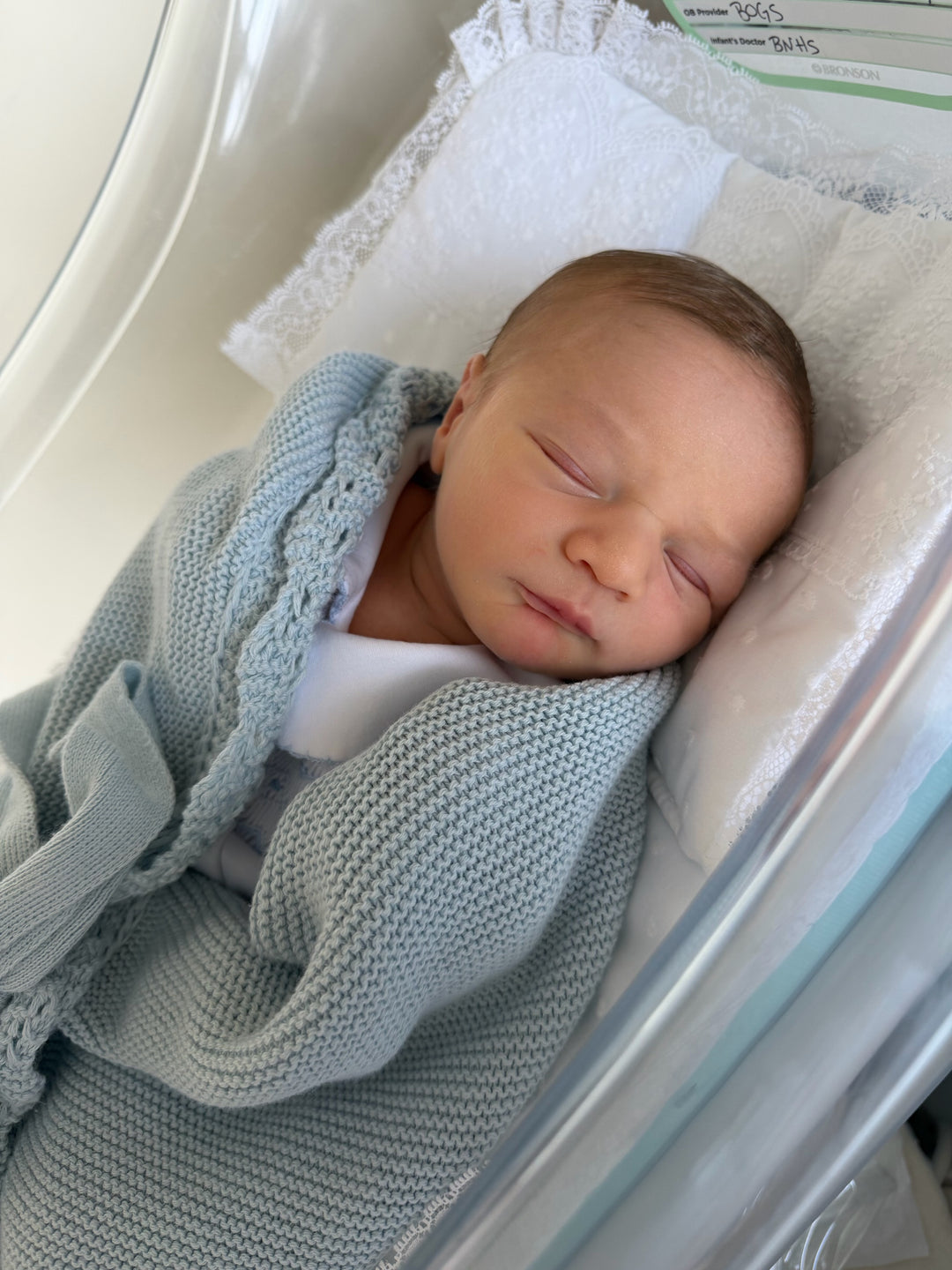 A newborn baby sleeping peacefully in a clear baby incubator, swaddled in a light blue knit blanket.