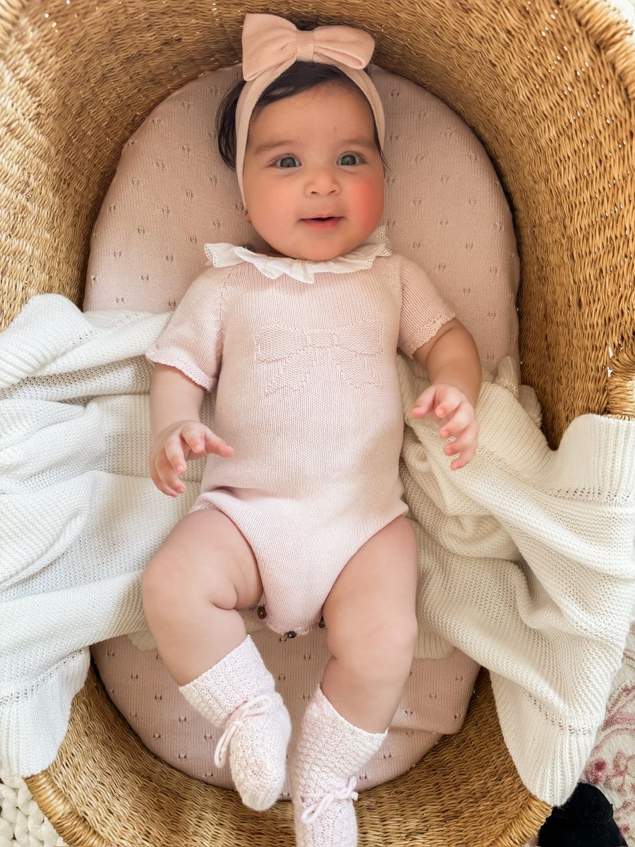 Baby in a pink outfit with a bow headband sitting in a wicker basket.