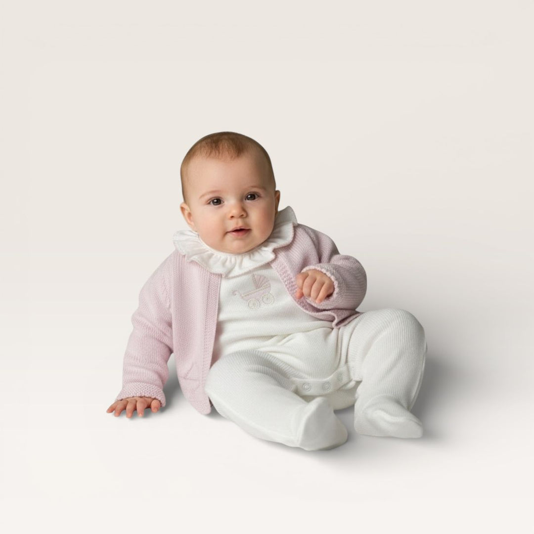A baby sitting on a light beige studio background, wearing a pale pink knit cardigan over a white ruffled collar top and white footed onesie.