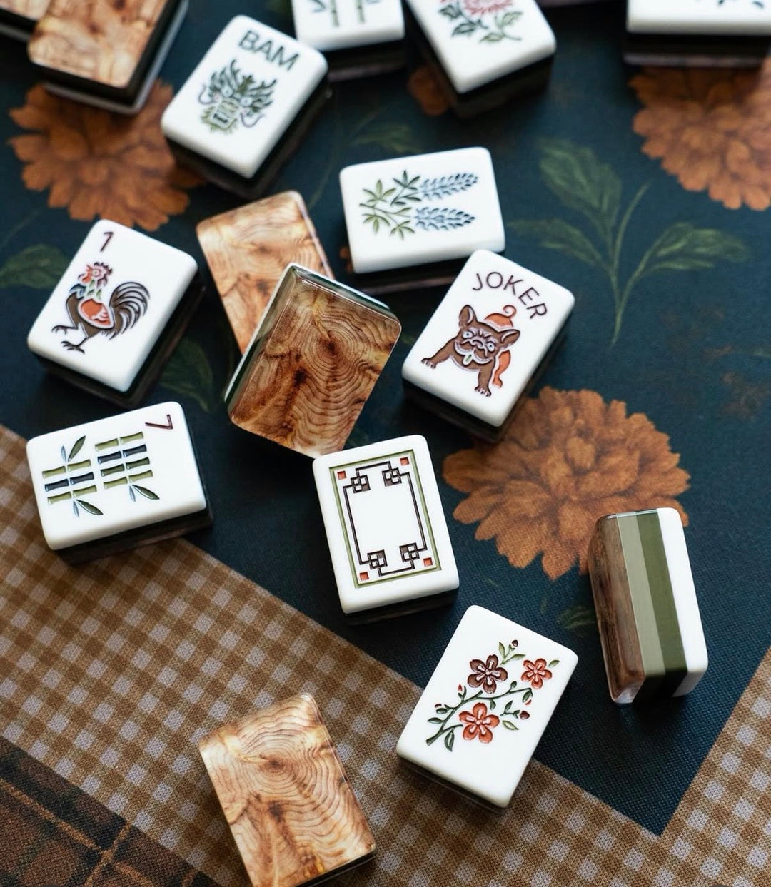 Mahjong tiles arranged on a floral and checkered surface, showing white tiles with printed illustrations and several wood-grain back tiles.