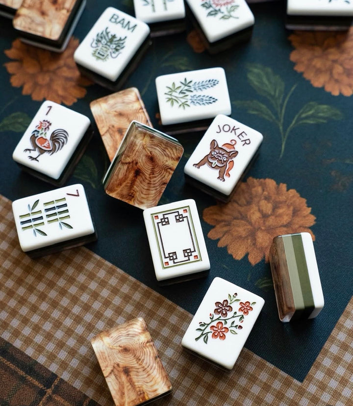 Mahjong tiles arranged on a floral and checkered surface, showing white tiles with printed illustrations and several wood-grain back tiles.