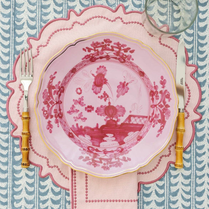 Decorative pink plate with floral design on a matching placemat, fork, and knife on a blue and white patterned tablecloth.