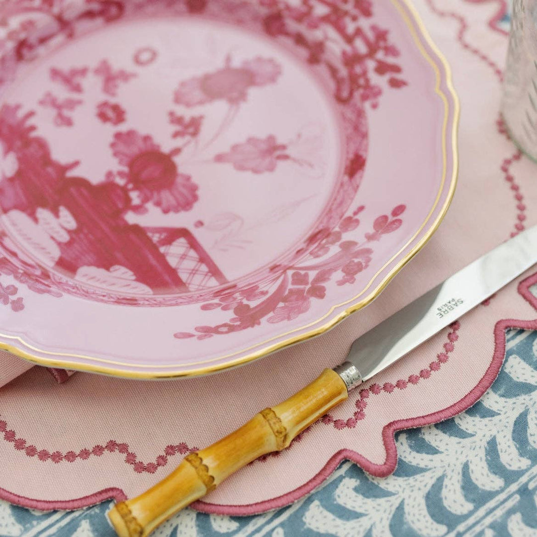 Detail view of a pink embroidered scalloped placemat with a decorative plate and bamboo flatware