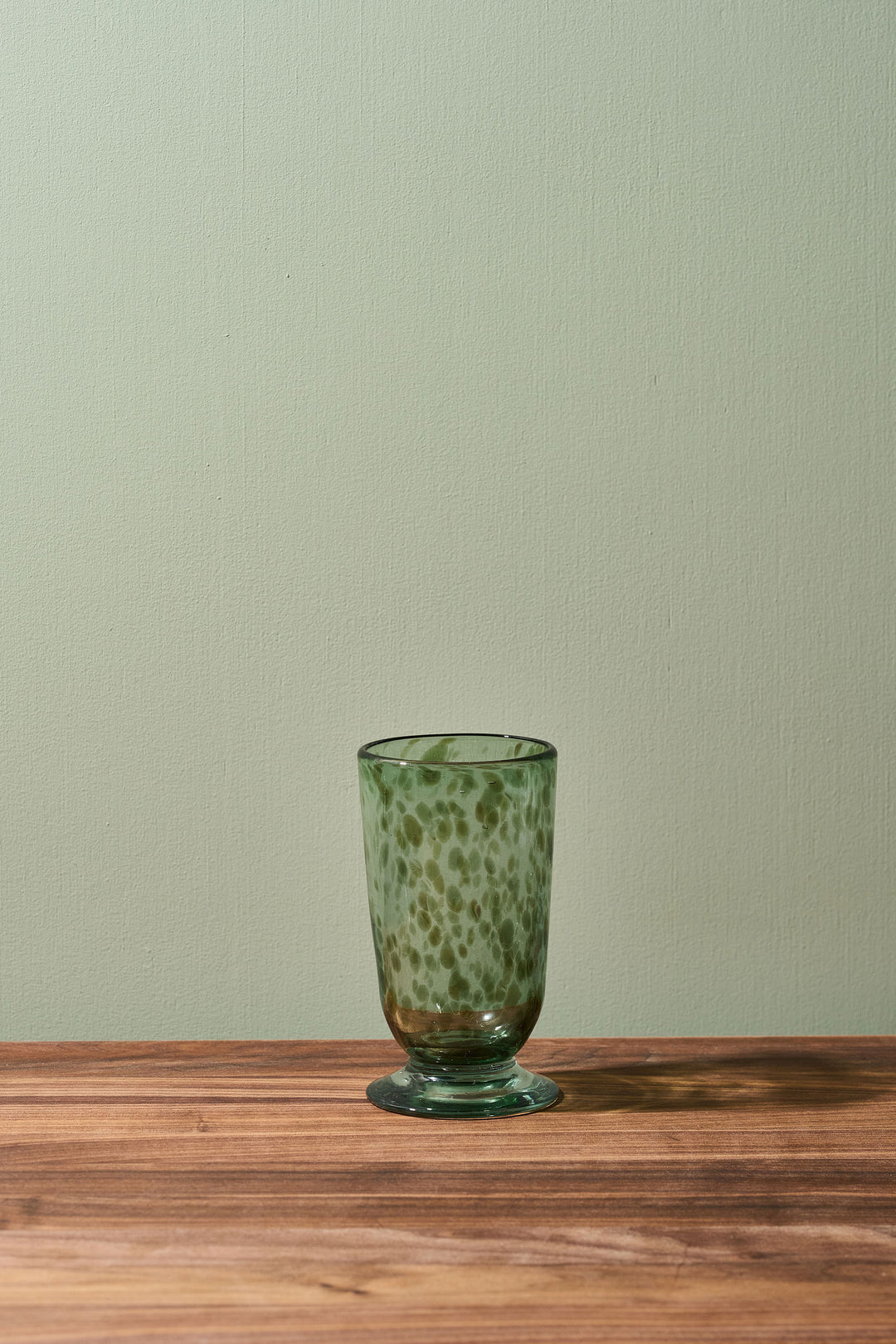 A small green glass tumbler with a speckled pattern and a circular base, set on a wooden surface against a light sage green wall.